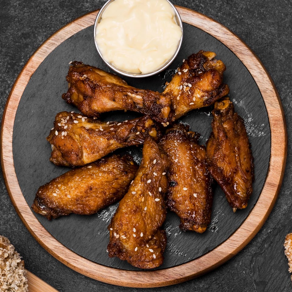 Six glazed chicken wings with sesame seeds and a side of creamy dipping sauce on a dark slate plate.