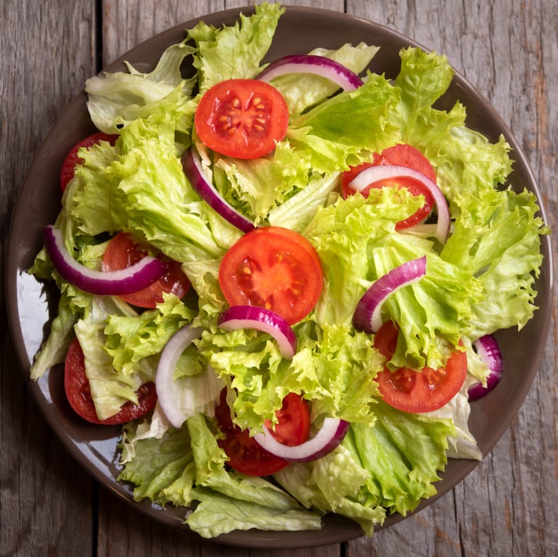 Fresh green lettuce, sliced red tomatoes, and purple onion rings in a brown bowl.