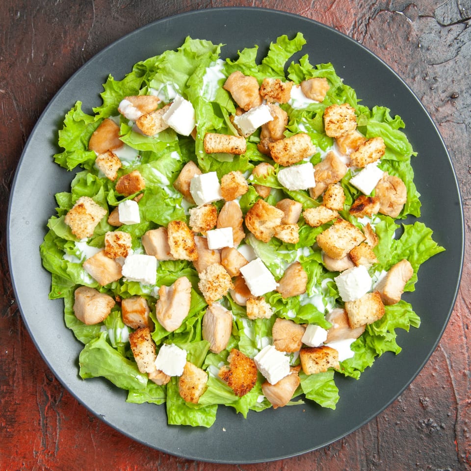 A Caesar salad with chicken, croutons, and feta cheese on a dark plate.